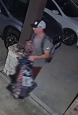 Camera footage photo of the first suspect wearing a balck and white ball cap, gray t-shirt and shoes and maroon shorts. This suspect is pictured carrying a gray backpack and a set of golf clubs.